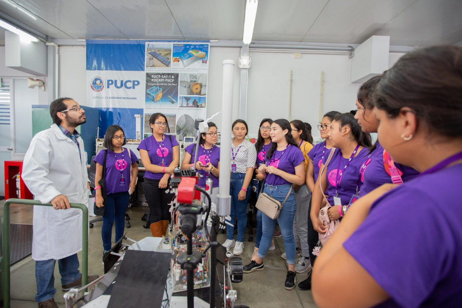 Un grupo de 10 alumnas están en un laboratorio PUCP escuchando la explicación del expositor.
