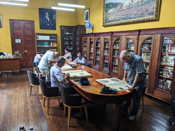 Tradicional sala de lectura de la biblioteca del IRA. Es una toma panorámica. En una mesa larga de madera hay 7 investigadores. El piso es de madera. Detrás, estantes de madera con libros hasta una altura aproximada de metro ochenta. Encima de ellos, las pareces, de techos altos, tienen cuadros.