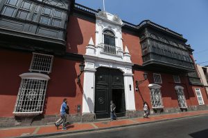 Fotografía de la casa Riva Agüero, csontruida a fines del sigo XVIII. Tiene dos pisos, techos altos, paredes rojas, balcones cerrados de madera labrada en el segundo piso y, al centro, una entrada en columnas blancas con un portón de madera oscura y grande.