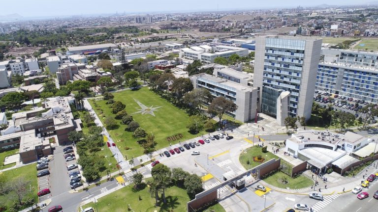 Toma aérea de la entrada principal de la PUCP. En el centro destaca un amplio jardín rectangular cuyo borde está rodeado de árboles y, a la derecha, el edificio mac gregor de 12 pisos, plomo con ventanas azules