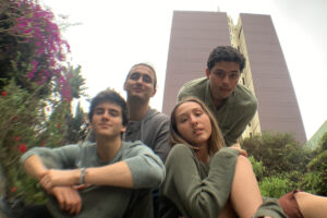 Cuatro jóvenes sentados viendo a la cámara. En primera lína un chico y una chica sentados en el pasto, ella apoya la espalda en el hombro de él. detrás de ambos, dos jóvenes se asoman por encima de ellos hacia la cámara. Están rodeados de arbustos, flores a la izquierda y, detrás, sobre salen dos edificios gemelos, torres grises.
