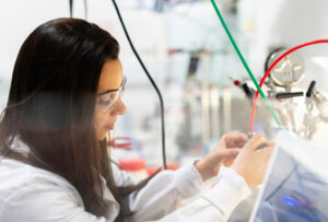 Ingeniera joven con una bata blanca. Se le ve de perfil, conectando una manguera delgada a alguún dispositivo. Lleva el pelo largo y lentes plásticos protectores