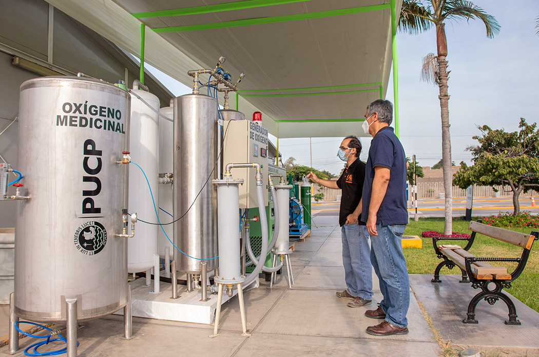 Plano abierto en el que se ve, del lado izquierdo, una planta de producción de oxígeno y tanques metálicos, el primero y más grande de ellos dice PUCP y tiene el logo en letras negras. Del lado derecho, dos hombres con mascarillas inspeccionan los tanques.