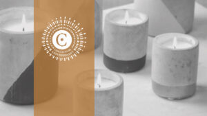 Velas gruesas encendidas en una foto a blanco y negro cálidamente iluminada. Hay una franja marrón hacia el lado izquierdo con el logo del Consorcio de UNiversidades: Una "C" marrón sobre fondo blanco