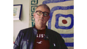 Hombre blanco de cabello muy corto, ligeramente calvo. Tiene lentes de marco grueso negro- Lleva un polo guinda y una casaca negra encima