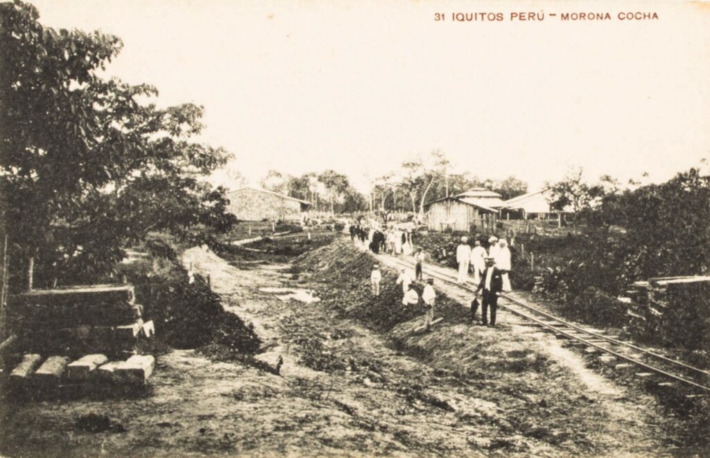 Foto en blanco y negro, toma abierta, se ve la ocnstrucción de vías de ferrocarril en medio de la selva y se lee "iquitos perú - morona cocha"