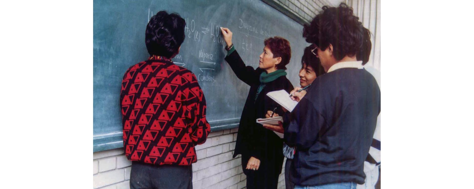 Tres alumnos con una profesora haciendo un repaso frente a una pizarra.