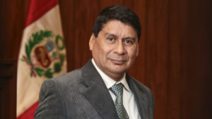 Retrato de plano medio del profesor willy carrera. Está con terno plomo, corbata a juego y camisa blanca. lleva el pelo corto y sonríe ligeramente. Detrás, desenfocada, l abandera del perú.