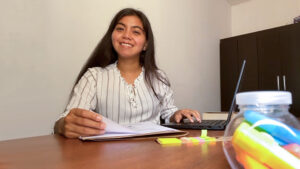 La egresada de Geografía y Medio Ambiente Katherine Lopez en su escritorio de trabajo. Viste blusa blanca con rayas oscuras.