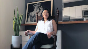 mariana chu en una silla en su sala. Usa blusa blanca sin mangas y pantalón oscuro. de fondo hay un cuadro