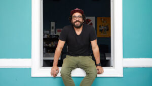 Fotografía de joven sentado sobre el marco una ventana abierta viendo a la cámara. El fondo es de un celeste encentido. Él lleva polo negro, gorra guinda, barba y lentes. Sonríe ampliamente