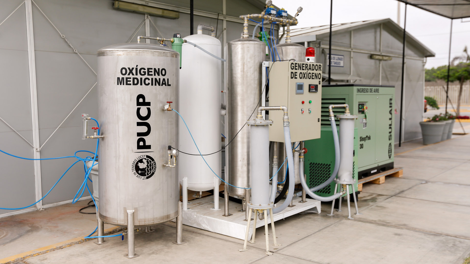 Fotografía de una planta de oxígeno PUCP. Al aire libre, se muestran un conjunto de componentes que conectan tanques y cajas metálicas, una de ellas el compresor y el centro de comando.