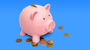 Fotografía de una alcancía con forma de chanchito rosado sobre fondo azul claro. Es tierno y caricaturesco. Tiene monedas alrededor.