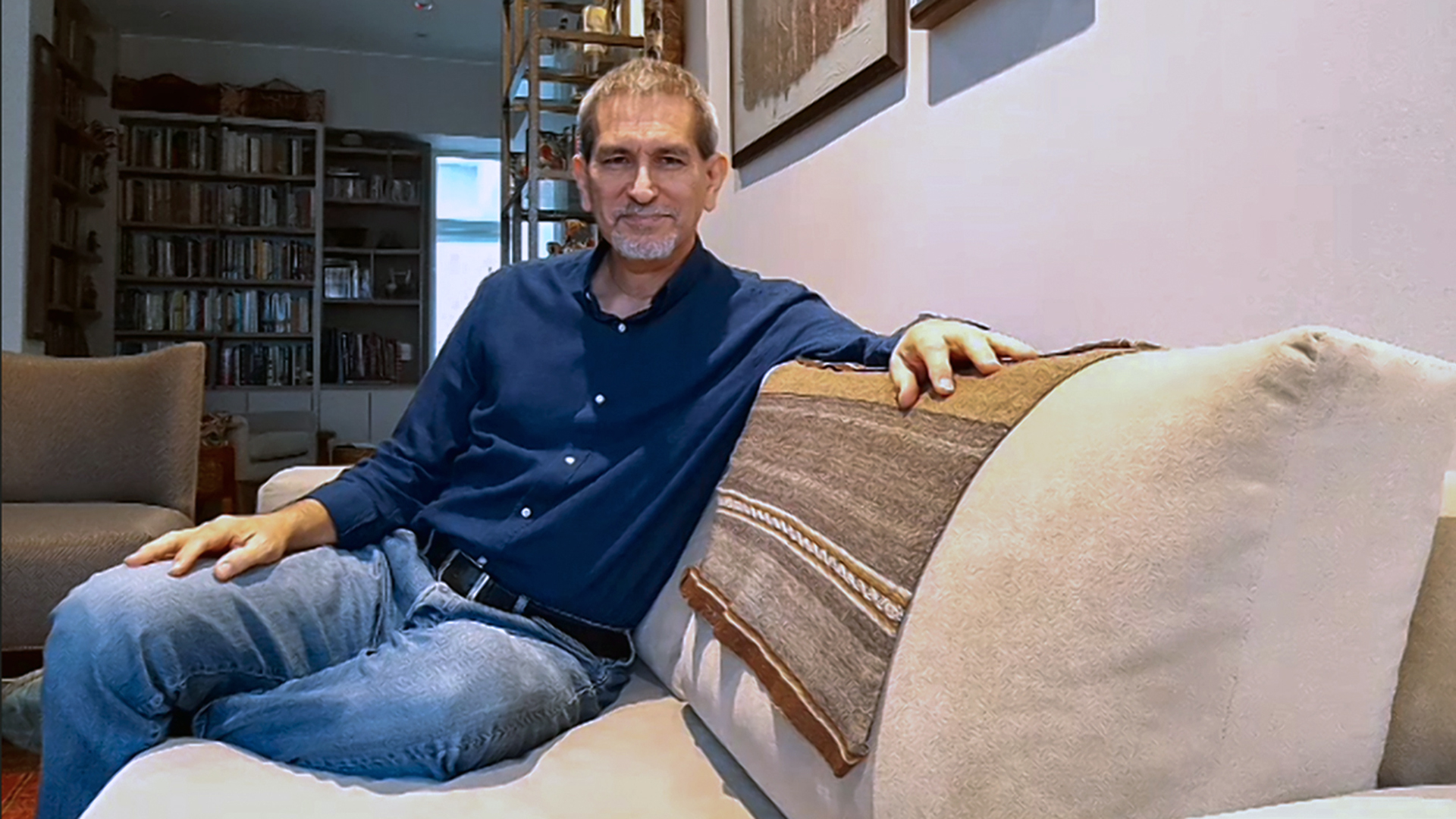 Raul renato Romero sentado en el mueble de su sala. Viste una camisa azul y jeans. En el fondo de ve su biblioteca.