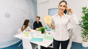Fotografía en la que se ve a dos personas trabajando en un escritorio y, en primer plano, una mujer con blusa y jean sotiene un foco de cartón mientras sonríe a la cámara.