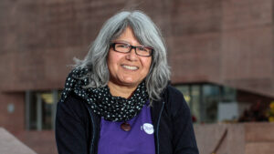 Foto de la profesora Pajuelo. Es una mujer de aproximadamente 50 años, de copmlexión robusta y cabello cano hasta los hombros. Lleva lentes de marco grueso y sonríe a la cámara. Tiene un polo morado y casaca y bufnada negra.