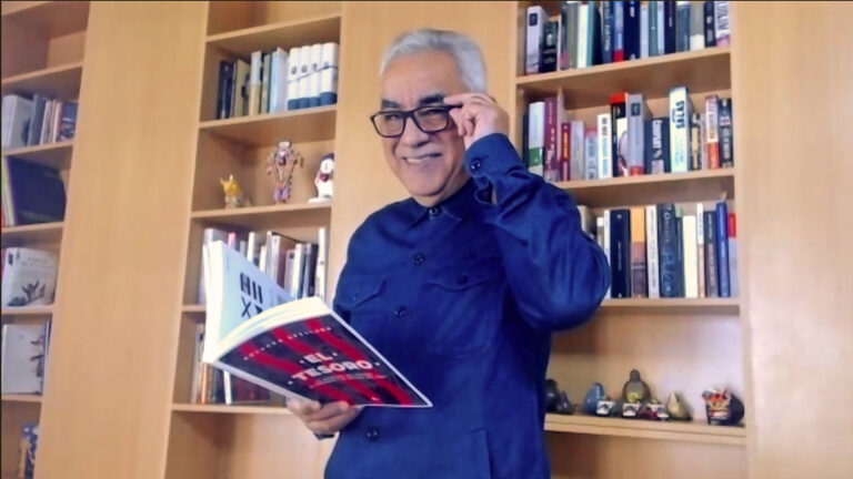 Rolando Arellano con camisa azul sosteniendo sus gafas puestas y su libro abierto en la otra mano
