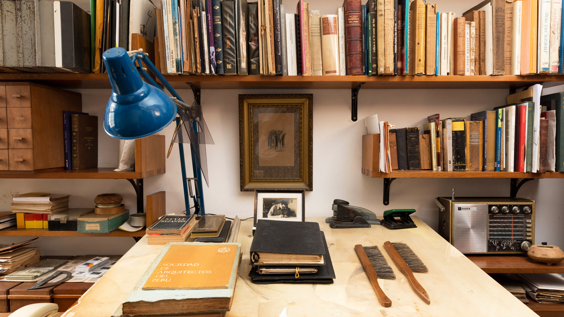 Foto moderna (la única de la serie) en la que se ve un escritorio con varios libros al frente y una lámpara azul que se puede desplazar sobre el escritorio.