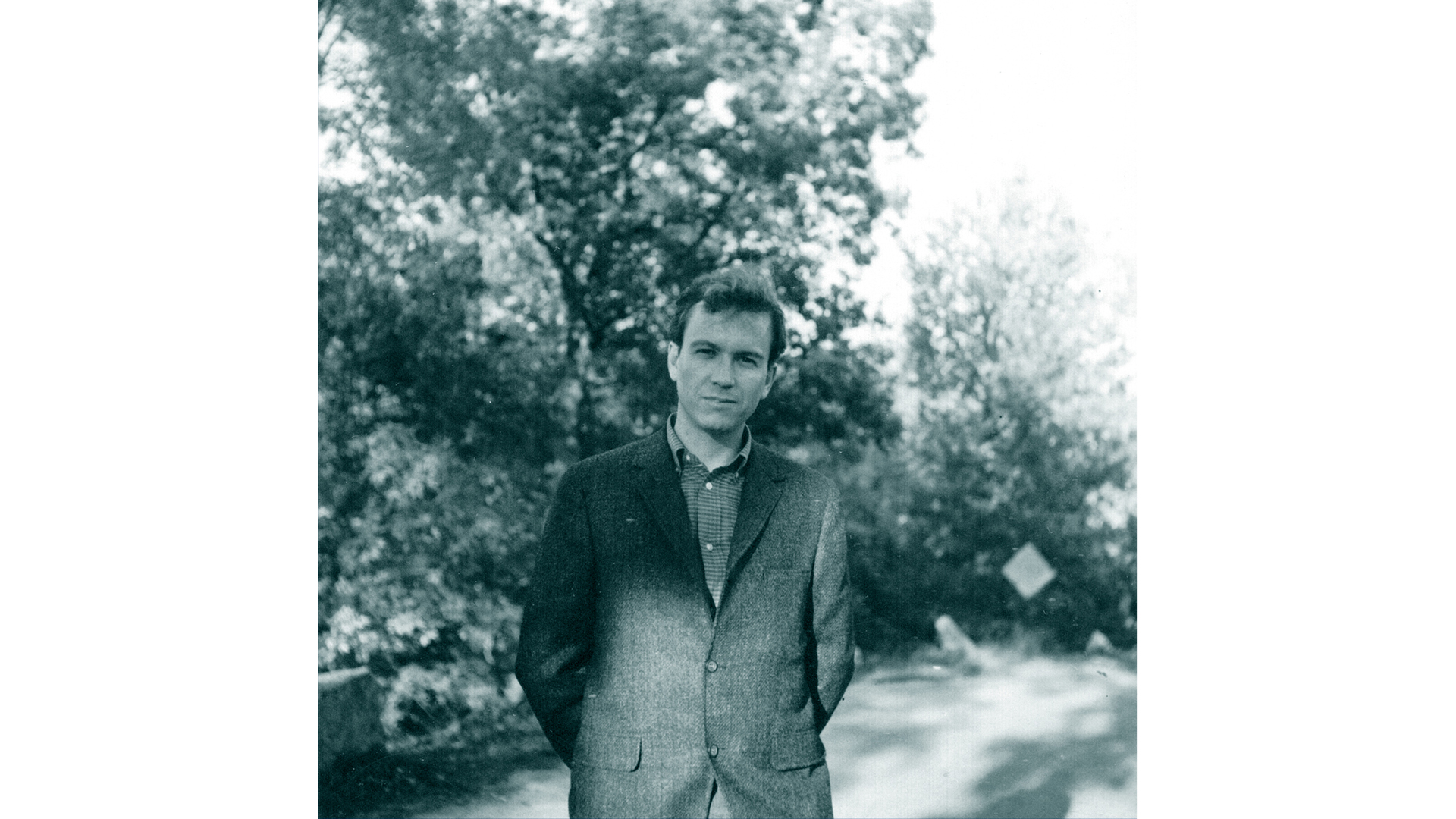 FOto en blanco y negro de tonos verdosos en la que aparece un señor, de cintura para arriba, con árboles en el fondo. el señor lleva un polo y saco cerrado encima.