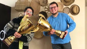 En la imagen, José Vergara y Mauricio Jarufe sonríen mientras sostienen sus trofeos. Son dos copas doradas de aproximadamente 60 cms de alto, cada una. José está a la izquierda, usa lentes y una chompa negra; Mauricio a la derecha también usa lentes y tiene un camisón azulino. De fondo una pared color crema en la que cuelgan cestos, sombreros y artesanías de paja.