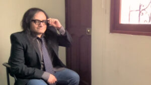 Richard Parra sentado en su estudio y con la mano izquierda toca sus anteojos. Usa el pelo largo y suelto.