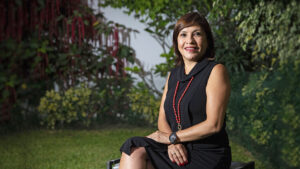Fotografía de Zamudio sentada en una banca de un jardín. se le ve desde las piertas hasta encima de la cabeza. Sonríe viendo a la cámara. Lleva el cabello marrón liso un poco por encima de los hombros con raya al costado derecho, labial rojo a juego con un collar grande, y un vestido negro sin mangas.