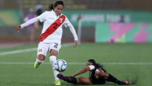 Foto de un partido de fútbol. En la toma se ve a una deportista de la slección de perú (short blanco, polo de manga larga blanco con una franja roja que cruza desde su hombro izquierdo a la zona inferior derecha). Está superando a una rival que se barre para tratar de quitarle el balón.