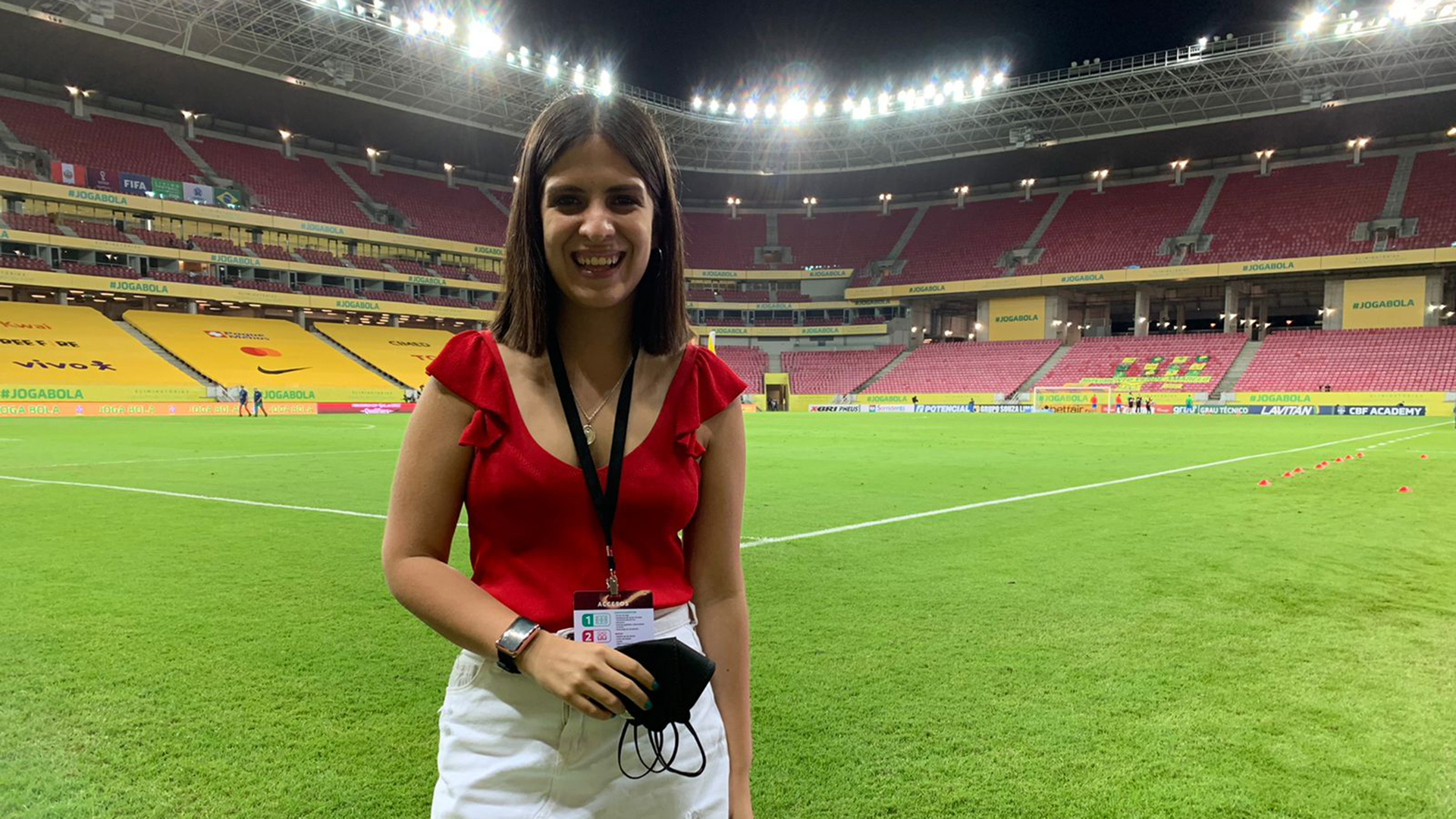 Foto amplia de una periodista con blusa roja y falda blanca en un estadio vacío, de noche, con las luces prendidas. la periodista sonríe ampliamente; lleva el pelo lacio con raya al medio y un brazalete dorado en la mano derecha.