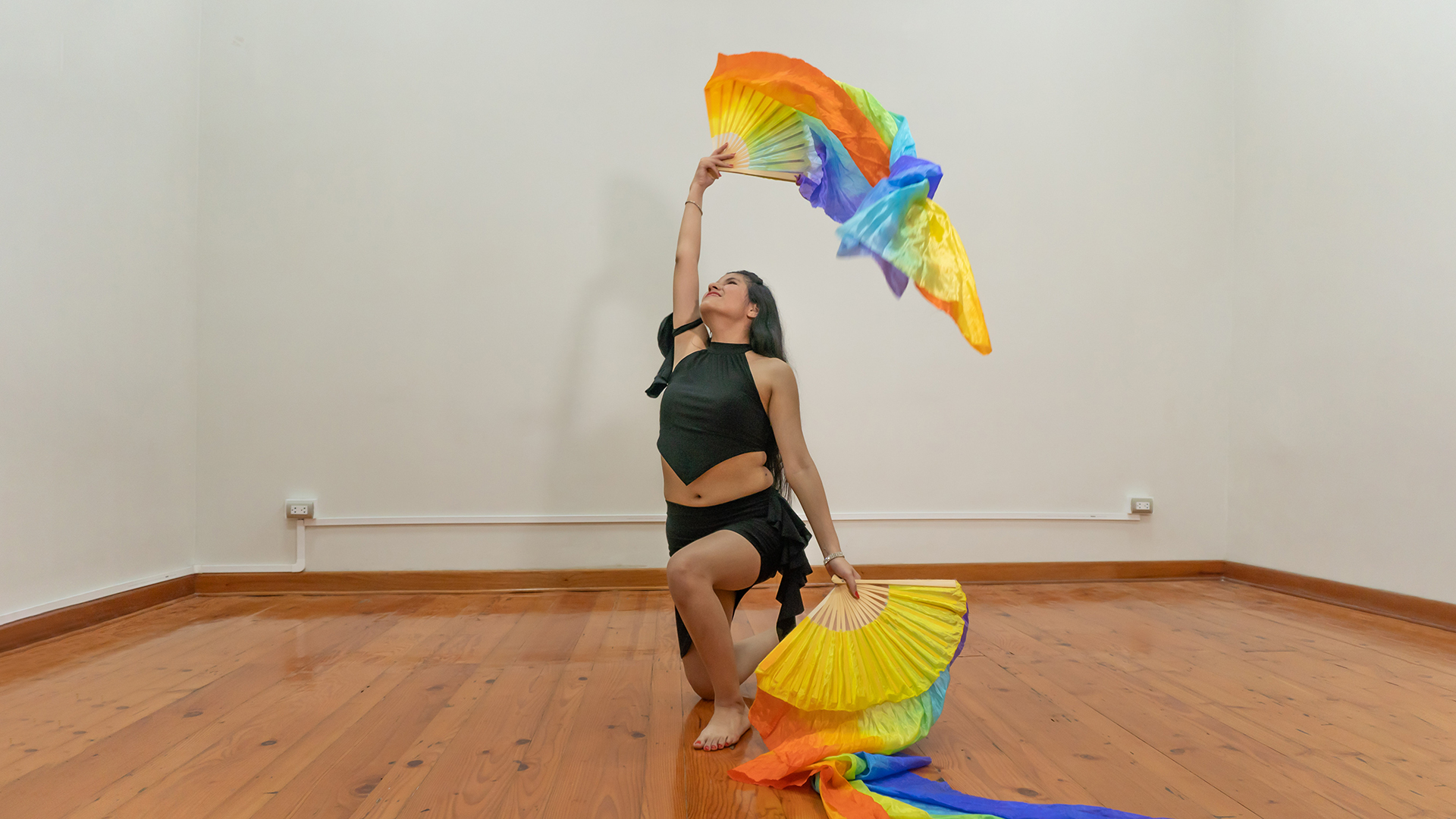 Andrea arrodillada sostiene un abanico en el piso y otro en el aire.