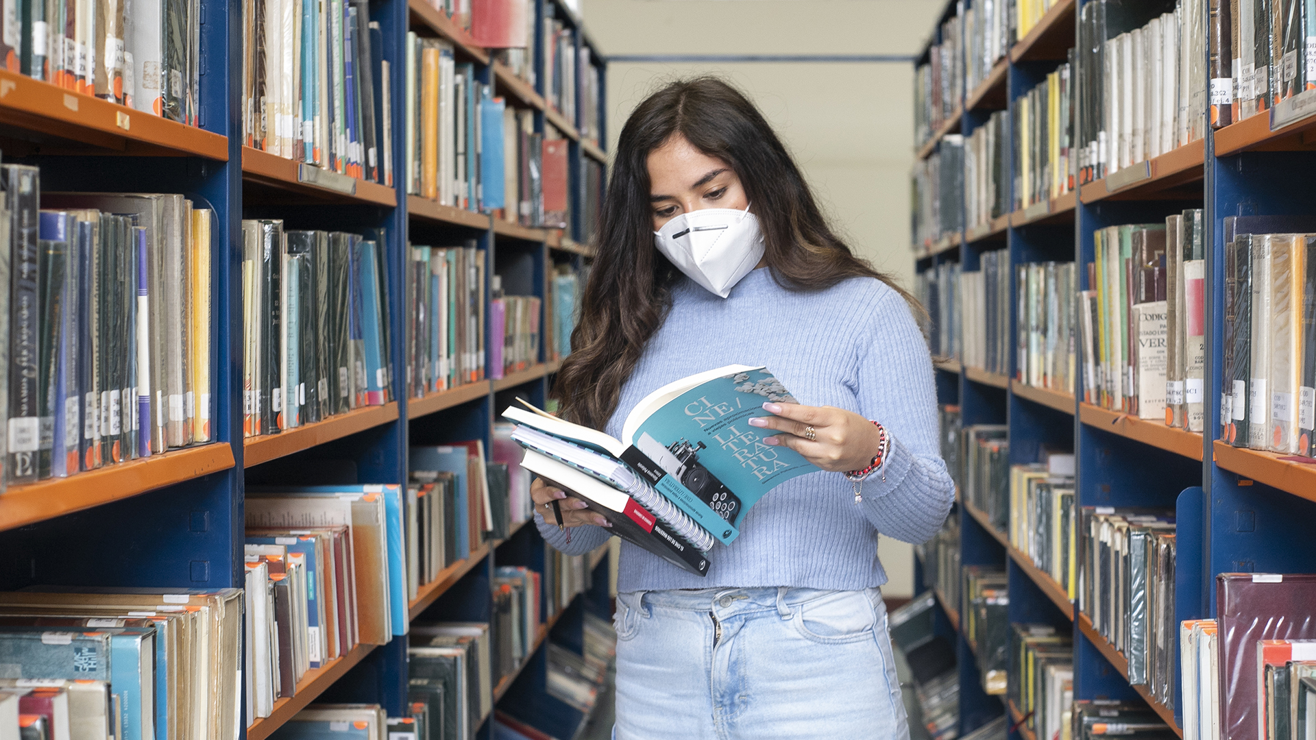 Foto de una estudiante de Becas PUCP. Está en la biblioteca con dos libros y un cuaderno. Refleja el esfuerzo para conseguir y mantener la Becas PUCP.