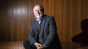 Retrato en foto del profesor emérito PUCP Luis Peirano. Es un señor de tez blanca de aprox 70 años con barba blanca y una ligera calvicie. Sonríe a la cámara. Lleva una camisa celeste y casaca y pantalón oscuros.