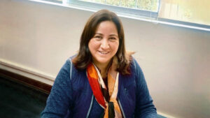 Foto de Ursula Zevallos, una mujer de aprox 40 años con cabello lacio que pasa de los hombros, saco azul y un pauelo largo rojo, amarillo y blanco. Sonríe a la cámara sentada en un escritorio.