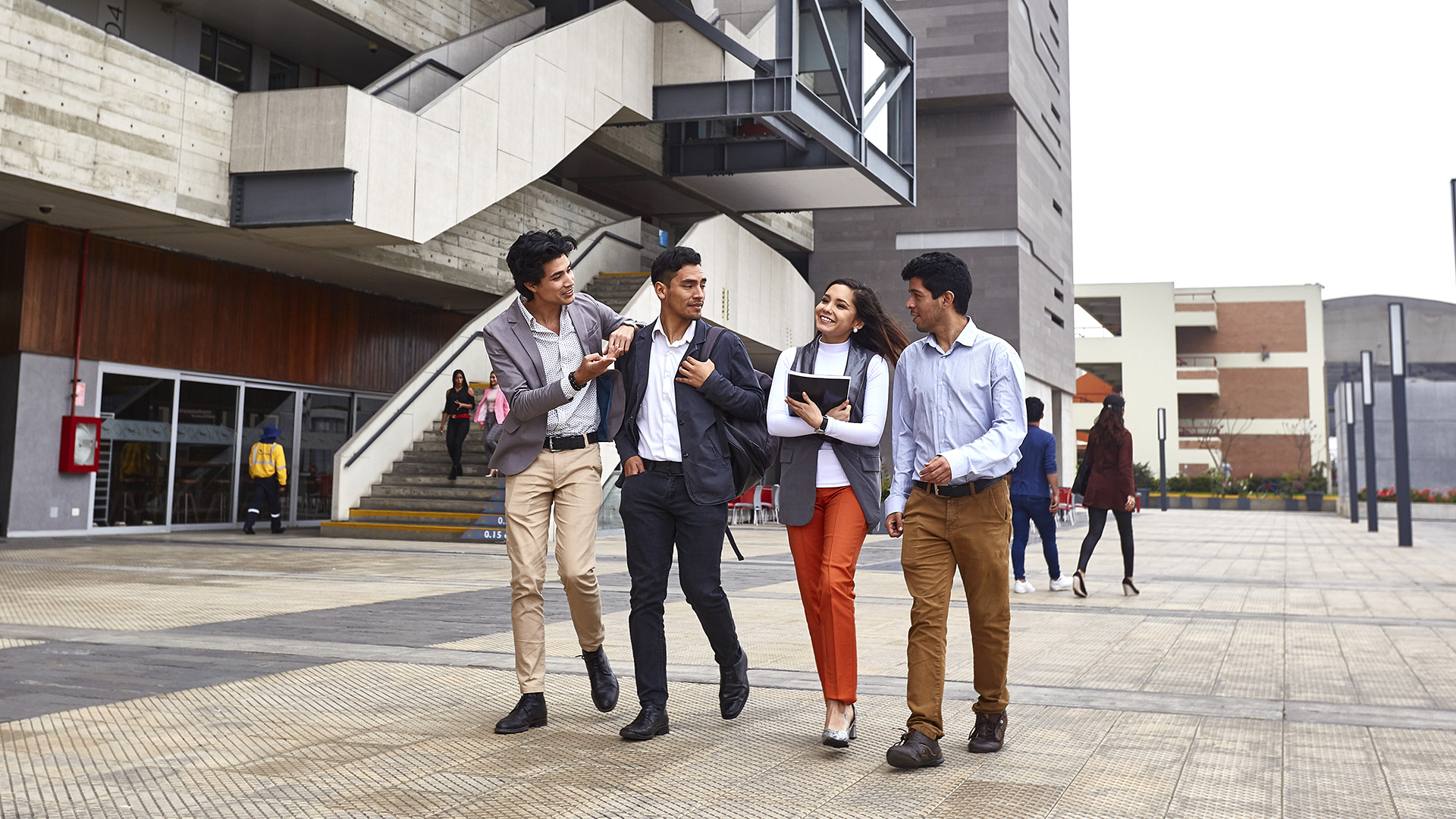 estudiantes de maestrias y doctorados de la escuela de posgrado pucp: cuatro estudiantes jóvenes de aproximadamente 30 años caminnado delante de un edificio moderno.