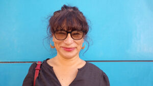 Foto de una mujer sonriente de aprox 40 años. Lleva el cabello oscuro recogido, cerquillo y lentes de sol con lunas amarillas. La pared de color celeste encendido contrasta con su tez clara y una blusa sport holgada oscura.