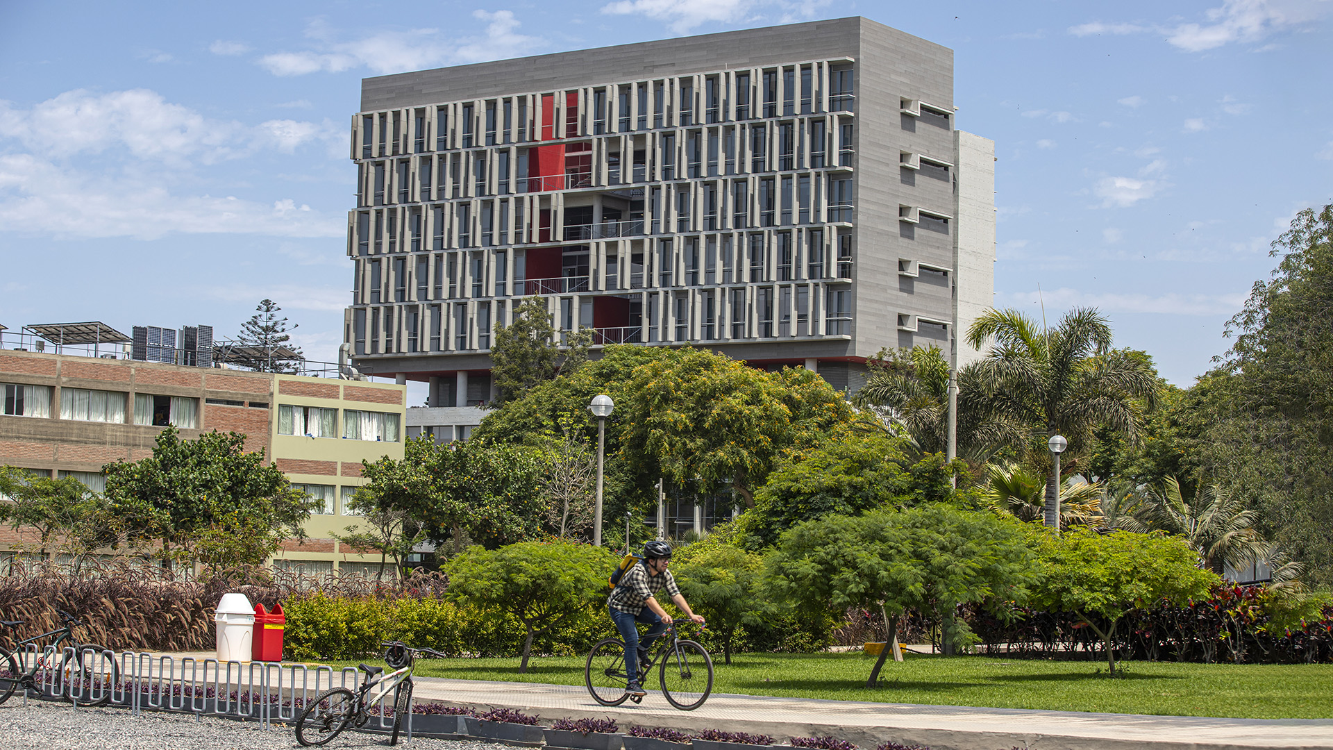 foto del campus de la PUCP: se ve un edificio de unos ocho pisos, más ancho que alto, en color plomo, ventanas con salientes y algunas aplicaciones en rojo. delante hay un jardín con árboles y en primer plano se ve un ciclista que pedalea en una vereda.