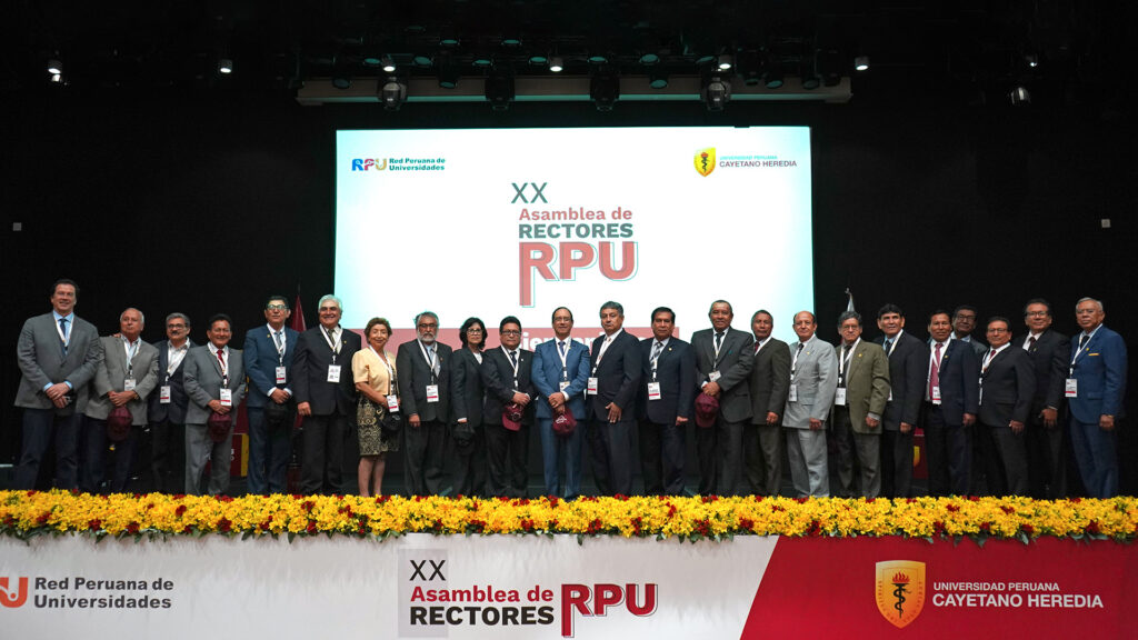 Foto de la asamblea de rectores RPU: 24 rectores, 22 hombres y 2 mujeres, parados en un estrado con el símbolo de RPU en el fondo