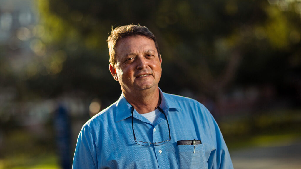 Foto del padre Fernando Roca ALcázar: un hombre de aprox 60 años con camisa celeste y lentes colgados del cuello. Sonríe a la cámara ligeramente.