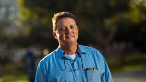 Foto del padre Fernando Roca ALcázar: un hombre de aprox 60 años con camisa celeste y lentes colgados del cuello. Sonríe a la cámara ligeramente.