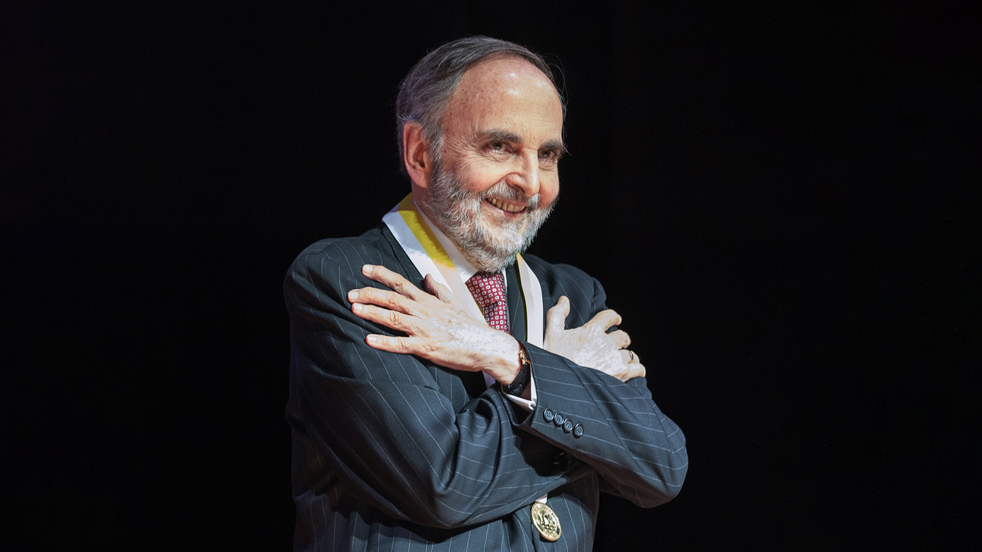 foto del profesor emérito javier de belaúnde lopez de romaña: un señor de aproximadamente 60 o más años, con terno, barba y una medalla. sonríe y hace un gesto de abrazo.