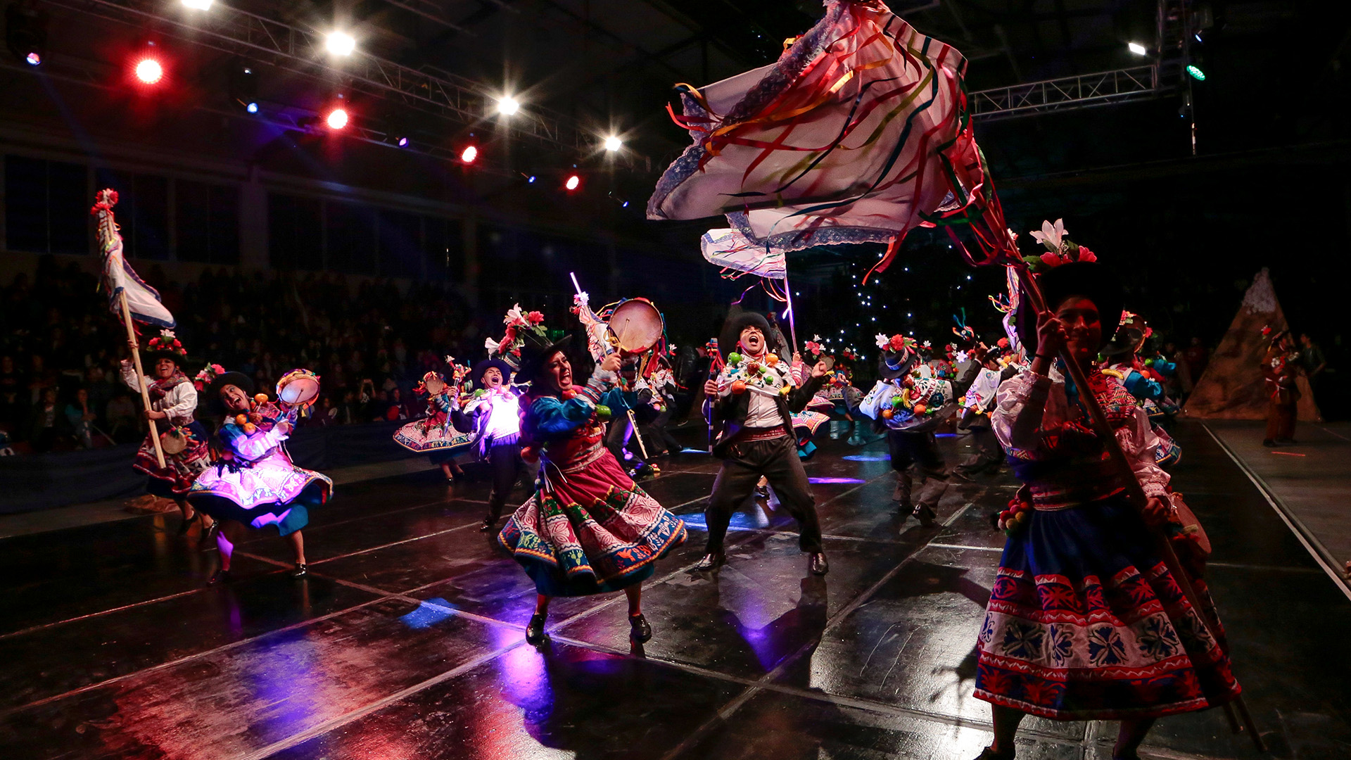 Noche cultural de los juegos interfacultades de la PUCP. En un escenario iluminado con luces moradas, unos 15 estudiantes bailan una danza folclórica.