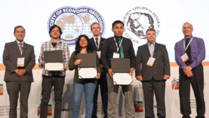 estudiantes PUCP que ganaron premios en proexplo: 3 jóvenes (2 hombres y una mujer) muestran diplomas de reconocimiento, rodeados de 4 señores vestidos con terno, De fondo se ven logos con el mundo sobre fondo blanco.