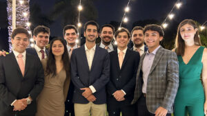 estudiantes PUCP destacan en torneo de debate en Harvard: grupo de 10 estudiantes, 8 hombres y 2 mujeres, vestidos de manera formal y sonriendo a la cámara. es de noche y de fondo se ven varios focos de luz amarilla. Todos los y las estudiantes sonríen y están alegres.
