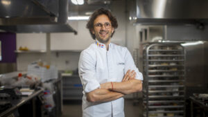 Jordi Bordas: una fotografía le muestra de cintura par arriba con camisa blanca de cocinero, remangada sobre los codos. es un hombre espigado con cabello castaño oscuro de largo mediano. tiene ojos que destacan, lentes de montura oscura, barba d epocos días y sonríe a la cámara con los brazos cruzados.