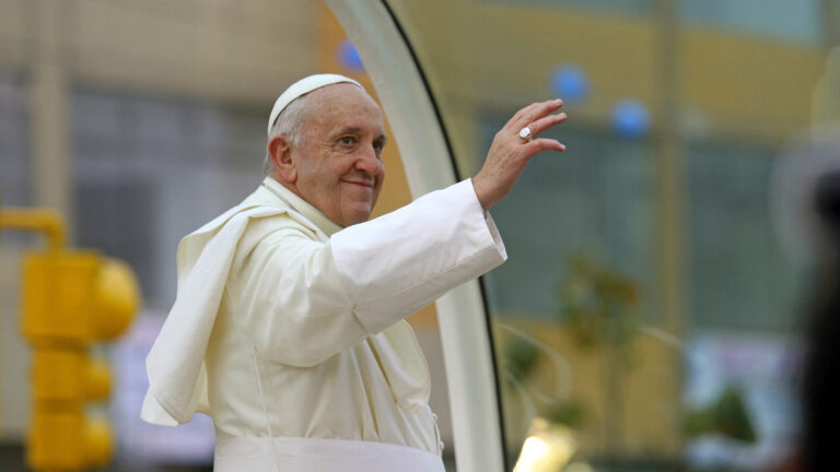 Foto del Papa Francisco saludando desde el papamóvil: un señor de más de 70 años, vestido de blanco con el atuendo de papa y una sortija grande.