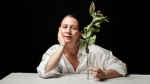 Foto de Rosario Yori: una mujer de aprox 40 años de tez clara, camisa blanca y el cabello rubio amarrado hacia atrás. Tiene los dos codos sobre una mesa y apoya la cabeza sobre su mano derecha. Con la mano izquierda sostiene una rama de hojas verdes que se están secando. Mira a la cámara con expresión neutra. Lleva los labios pintados de rojo.