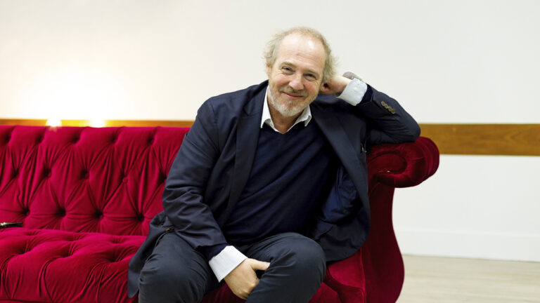 Foto de Arnaud Desplechin sentado sobre un sofá rojo sobre fondo blanco. Es un señor de aprox 60 años con traje oscuro y chompa azul. Lleva el pelo cano suelto y barba cana de pocos días.