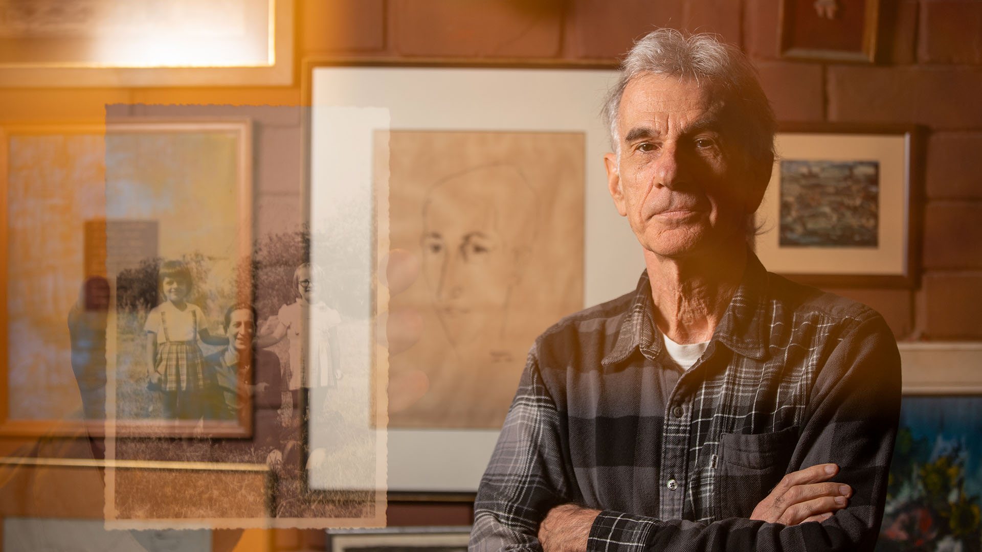 Hombre en camisa de cuadros, con los brazo cruzados frente a ilustraciones y cuadros
